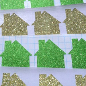 May include: A sheet of green and gold glitter paper cut into house shapes. The houses are arranged in rows on a white grid background.