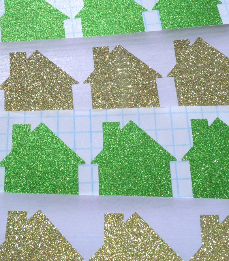 May include: A sheet of green and gold glitter paper cut into house shapes. The houses are arranged in rows on a white grid background.