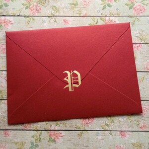 Könnte beinhalten: Ein roter Umschlag mit einem goldenen Buchstaben "P" in der Mitte. Der Umschlag besteht aus strukturiertem Papier und ist versiegelt. Der Hintergrund ist ein Blumenmuster auf einer Holzoberfläche.