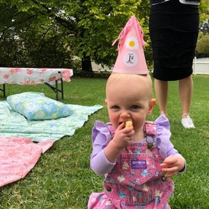Puede incluir: Un ni&ntilde;o peque&ntilde;o con un peto floral rosa y una camisa morada de manga larga. El ni&ntilde;o lleva un gorro de cumplea&ntilde;os rosa con una margarita y el n&uacute;mero 1. El ni&ntilde;o est&aacute; sentado en la hierba y comiendo un bocadillo.