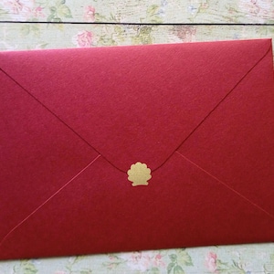 Könnte beinhalten: Ein roter Umschlag mit einem goldenen Muschel-Siegel. Der Umschlag besteht aus strukturiertem Papier und ist mit einem goldenen Aufkleber versiegelt. Der Hintergrund ist eine helle Holzoberfläche mit floralen Akzenten.