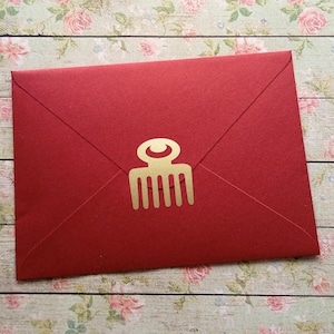 Könnte beinhalten: Ein roter Umschlag mit einem goldenen kammförmigen Emblem. Der Umschlag ist versiegelt und hat ein schlichtes, elegantes Design. Der Hintergrund ist ein Blumenmuster auf einer Holzoberfläche.