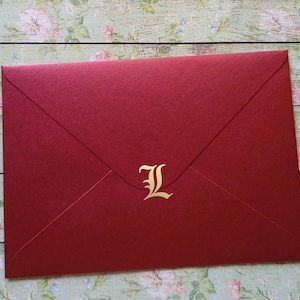 Può includere: Una busta rosso scuro con una lettera "L" dorata nell'angolo in basso a destra. La busta è chiusa e ha una classica patta a punta. Lo sfondo è un motivo floreale su una superficie di legno.