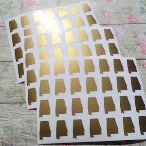 May include: Sheets of gold-colored Alabama state stickers. Each sheet contains multiple stickers with the state's outline. The stickers are on a white background, with a floral pattern visible in the background.