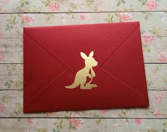 May include: A red envelope with a gold kangaroo silhouette on the front. The envelope is closed and has a textured surface. The background is a floral pattern on a wooden surface.
