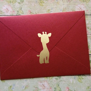 Può includere: Una busta rosso scuro con una silhouette dorata di giraffa. La busta è realizzata in carta strutturata e ha una classica chiusura a patta triangolare. La giraffa è centrata sul davanti.