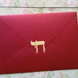 Op de afbeelding: Een rechthoekige rode envelop met een goudkleurig Hebreeuws Chai-symbool in het midden van de klep. De envelop heeft een puntige driehoekige sluitklep en een gladde, matte textuur, met een egale dieprode kleur over de gehele buitenkant.