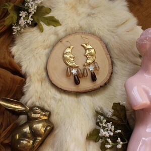 May include: Gold-colored crescent moon earrings with dangling brown and clear beads and white star charms. The earrings rest on a wooden disc, displayed on a fluffy, off-white surface. A brass rabbit figurine and pink statue are also visible.