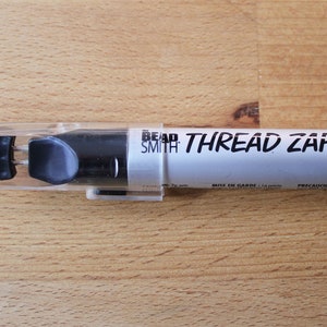 May include: A black and white Thread Zap II tool by Bead Smith, in a clear plastic sleeve. The tool has a black handle and a metal tip. The text "THREAD ZAP II" is printed on the white body of the tool.