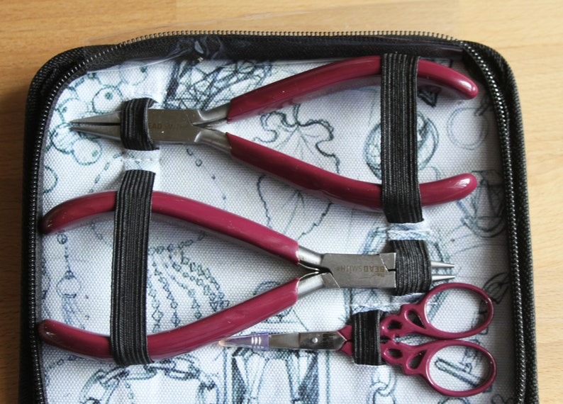 Puede incluir: Una funda negra con cremallera con un interior de tela blanca que contiene tres herramientas para hacer joyas: un par de alicates de mango rojo, un par de alicates de mango plateado y un par de tijeras de mango rojo.