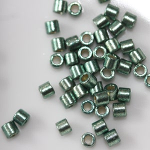 May include: Small, cylindrical beads in a metallic teal hue. Each bead has a central hole, scattered on a white surface. These beads are ideal for jewellery making and various craft projects. Each bead is approximately 3 mm in length.
