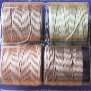 May include: Six spools of thread in a plastic pack. The spools are arranged in two rows of three. The thread colours are beige, light brown, and cream.