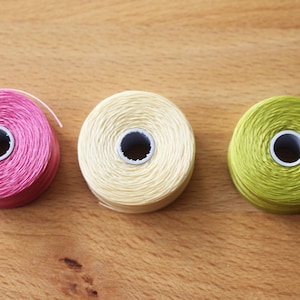 Könnte beinhalten: Drei Garnrollen, eine rosa, eine cremefarben und eine grüne, sind in einer Reihe auf einer Holzoberfläche angeordnet.