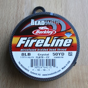 Könnte beinhalten: Eine Spule mit 8 lb FireLine microfused geflochtenem Perlenfaden in der Farbe Crystal. Die Spule ist 50 Yards lang und hat einen durchschnittlichen Durchmesser von 0,007 Zoll. Das Etikett enthält auch den Produktcode FLBT8-CY und den Barcode 7 90524 64115 4.