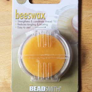 Könnte beinhalten: Eine Packung The Beadsmith Bienenwachs zum Stärken und Konditionieren von Fäden. Das runde, orangefarbene Bienenwachs befindet sich in einem transparenten Kunststoffbehälter. Der Packungstext lautet "beeswax" und "Strengthens & conditions thread".