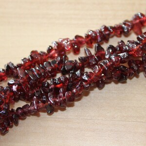 May include: Three strands of deep red garnet chip beads are shown against a light wood surface. The beads have a rough, irregular shape and a rich, translucent colour, suggesting a natural gemstone.