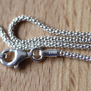 Könnte beinhalten: Nahaufnahme einer silbernen Halskette mit einer strukturierten, perlenbesetzten Kette. Der Karabinerverschluss ist sichtbar, mit der Aufschrift "ITALY". Die Halskette liegt auf einer hellbraunen Holzoberfläche.