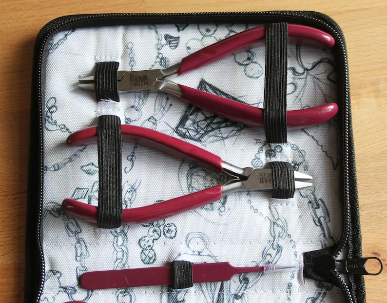 Puede incluir: Una funda negra con cremallera con un interior de tela blanca que contiene tres herramientas para hacer joyas. Las herramientas incluyen un par de alicates de mango rojo con el texto "Bead Smith" en el lateral, un segundo par de alicates de mango rojo y una peque&ntilde;a herramienta de mango rojo con una punta afilada.
