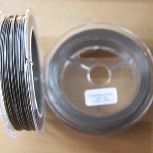 May include: Two spools of silver-coloured wire, each wound around a clear plastic spool. The spools are on a wooden surface.