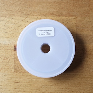 May include: A white spool of WrightWays Beads Tiger Tail, 0.35mm x 100m, is shown. The spool is circular with a central hole. The label has black text. The background is a light brown wooden surface.