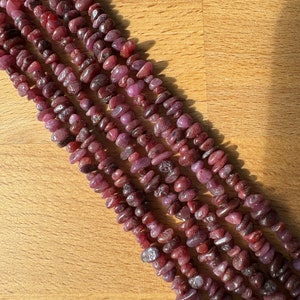 Könnte beinhalten: Eine Strähne aus chipsförmigen Rubinperlen in Rosatönen und Rot. Die Perlen sind in Reihen auf einer Holzoberfläche angeordnet.