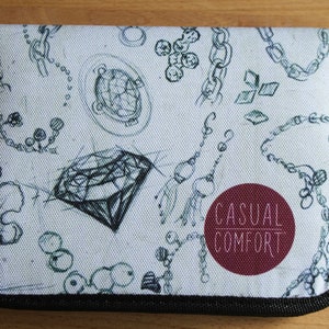 Puede incluir: Un estuche para l&aacute;pices de lona blanca con una cremallera negra y un borde negro. El estuche tiene un dise&ntilde;o en blanco y negro de cadenas, diamantes y otras joyas. El texto "Casual Comfort" est&aacute; impreso en blanco sobre un c&iacute;rculo granate.