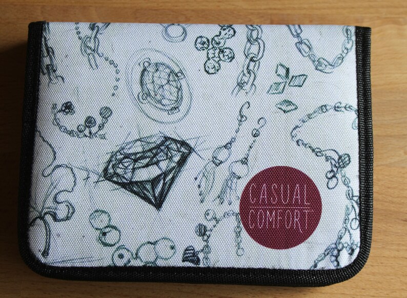 Puede incluir: Un estuche para l&aacute;pices de lona blanca con una cremallera negra y un borde negro. El estuche tiene un dise&ntilde;o en blanco y negro de cadenas, diamantes y otras joyas. El texto "Casual Comfort" est&aacute; impreso en blanco sobre un c&iacute;rculo granate.