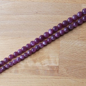 May include: Two strands of purple gemstone beads on a wooden surface. The beads are round and have a slight shimmer.