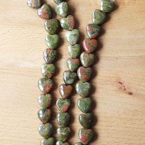 Könnte beinhalten: Eine Kette aus grünen und roten herzförmigen Perlen aus poliertem Stein. Die Perlen sind auf einem durchsichtigen Faden aufgefädelt und in einem Muster aus abwechselnden Farben angeordnet.