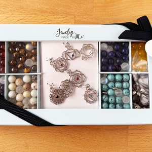 May include: A white gift box with a black ribbon and a heart-shaped tag that says "Chakra Bead Gift Set". The box contains seven compartments with various coloured beads and silver charms.