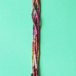 May include: A strand of multi-coloured gemstone beads in shades of pink, red, and orange. The beads are small and round, and they are strung on a clear thread. The beads are arranged in a graduated pattern, with the darkest beads at the top and the lightest beads at the bottom.