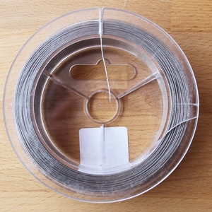 May include: A spool of silver-coloured wire in a clear plastic container. The wire is wound tightly around the spool.