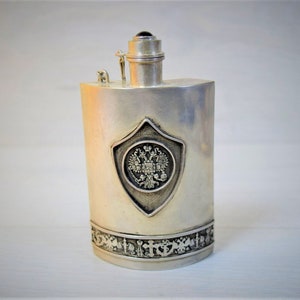 Peut inclure: Une gourde en argent avec un design orné représentant un blason avec un aigle à deux têtes. La gourde a un couvercle à charnière et une petite chaîne attachée au couvercle.