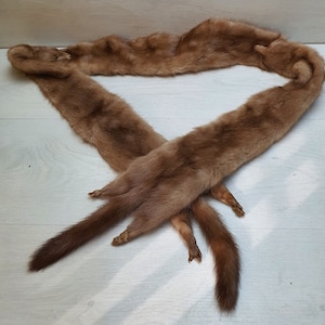 May include: A brown fur stole, possibly mink, lies on a light wood surface. The stole is long and narrow, with a soft, textured appearance. The tails of the animal are visible at the ends of the stole.