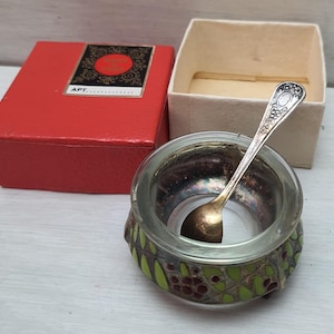 May include: A small glass salt cellar with a green and gold enamel design and a silver spoon. The salt cellar is in a red box with the word "APT" printed on it.