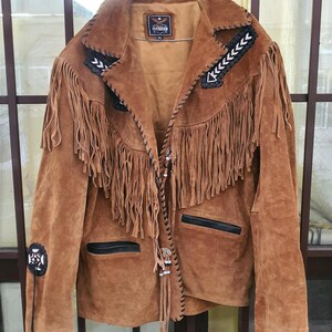 Puede incluir: Chaqueta de ante marrón con detalles de flecos y costuras negras. La chaqueta tiene cierre de cremallera y dos bolsillos. La etiqueta de la chaqueta dice "Vintage".