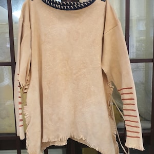 May include: A tan leather shirt with a black braided trim around the neck. The sleeves have red stripes and fringe along the edges.