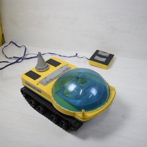 May include: A yellow toy rover with a blue dome and a gray antenna. The rover has a black track and is on a white surface. The rover is connected to a yellow remote control with two black buttons.
