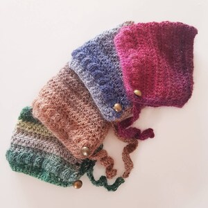 Op de afbeelding: Vier gebreide mutsen in verschillende kleuren, waaronder groen, bruin, blauw en roze. Elke muts heeft een decoratieve knoop.