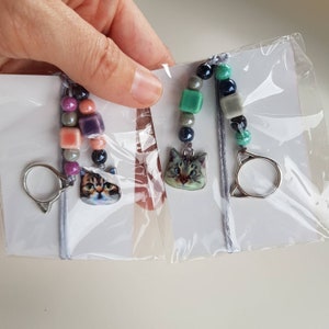 Puede incluir: Dos llaveros con temática de gatos con anillos plateados y cuentas de colores. Un llavero tiene cuentas rosas, moradas y grises, mientras que el otro tiene cuentas verdes, azules y grises.