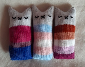 May include: Three knitted cat toys with different coloured stripes. The first cat is pink, purple and blue. The second cat is white, pink and blue. The third cat is brown, orange and purple.