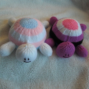 May include: Two knitted turtle toys. One is pink, white and blue with a light blue circle in the centre. The other is purple, white and black with a pink circle in the centre.