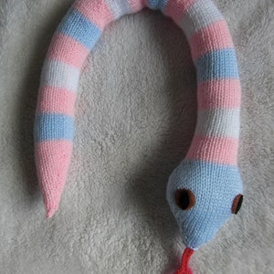 May include: A knitted snake toy with pink, blue and white stripes. The snake has a red tongue and brown eyes.