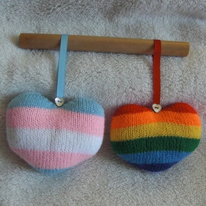 May include: Two knitted heart-shaped ornaments hanging from a wooden dowel. The heart on the left is pink, white, and light blue, while the heart on the right is rainbow coloured.