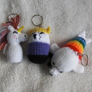 May include: Three knitted keychains: a unicorn with a pink and white mane, a bear with a yellow, white, purple and black striped body, and a white polar bear with a rainbow stripe on its back.