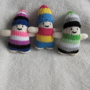 May include: Three knitted dolls with different coloured stripes and faces. The dolls are wearing hats with stripes in the same colours as their bodies. The dolls are sitting on a white fluffy surface.