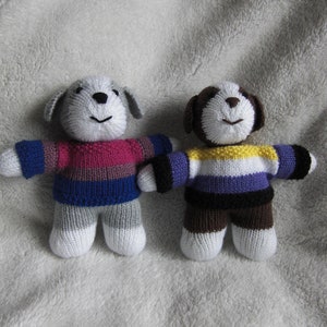 May include: Two knitted plush toys shaped like dogs wearing sweaters with the colours of the bisexual and asexual pride flags. The dog on the left is wearing a pink, blue and white sweater. The dog on the right is wearing a yellow, purple, black and white sweater.