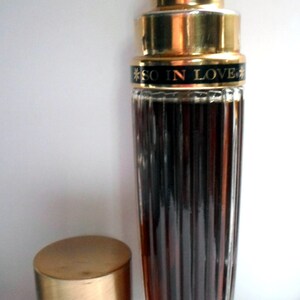 May include: A gold and clear glass bottle of perfume with a gold cap and a black label that reads "SO IN LOVE".