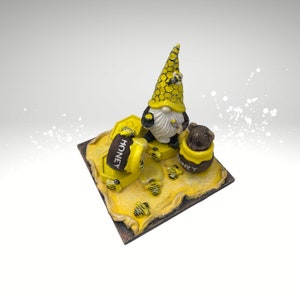 Könnte beinhalten: Eine gelbe und schwarze Gnomfigur, die einen Honigtopf mit Bienenmotiv hält. Der Gnom steht auf einem gelben und schwarzen Honigwabensockel mit Bienen und dem Wort "Honey" darauf.