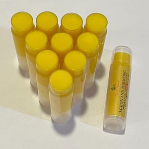 May include: A group of 10 yellow lip balm tubes with a single tube in the foreground. The tube in the foreground has a label that reads "Lemon and Turmeric Organic Moisturizing Lip Balm".